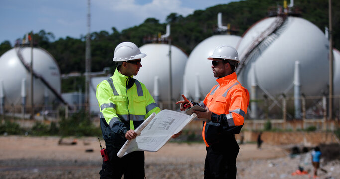 Two male power plant engineers work with a blueprint and a digital tablet by the seaside at a refinery and gas petrochemical plant