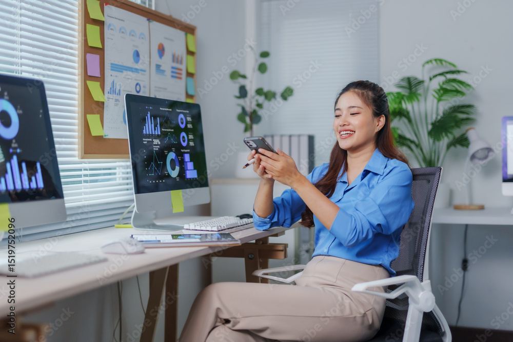 Fototapete young office worker engaging with a smartphone while analyzing graphs on a computer at a modern desk