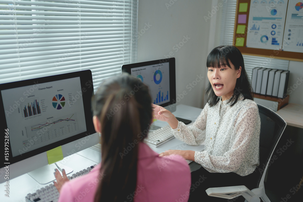 Poster two asian businesswomen discussing financial reports while working on a computer in a modern office 