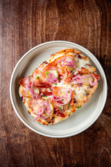 A delicious open-faced sandwich with melted cheese, crispy bacon, pickles, and pickled red onions, garnished with fresh parsley, served on a white dish atop a rustic wooden table.