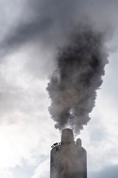 Vertical smoke stream from industrial pipe reaching cloudy skies, focus on contrast between dark emissions and bright background for climate topics.