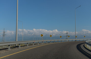 view of the bend of an empty highway with yellow road signs against a background of blue sky, clouds and mountains
