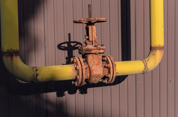 an old rusty valve on a yellow gas pipe against the background of a brown fence