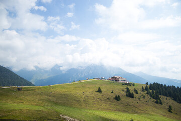 Mountain lodge on a green alpine ridge under a bright cloudy sky, surrounded by forest and peaks.