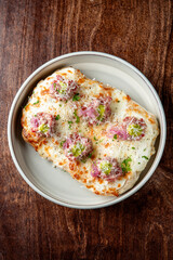 A close-up of a rectangular pizza with melted cheese, minced meat, pickled onions, and herbs, served in a round dish on a wooden table