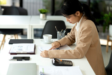 Young business woman is sitting at a desk working with documents charts and a calculator on the desk..