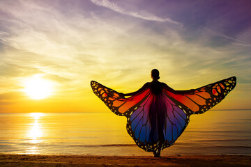 Graceful Woman with Vibrant Butterfly Wings on the Beach at Sunset Sunshine. Magical Mood of Transformation. Healing Meditation. Rear View of Girl Silhouette with Open Arms at Colorful Sea Sky view