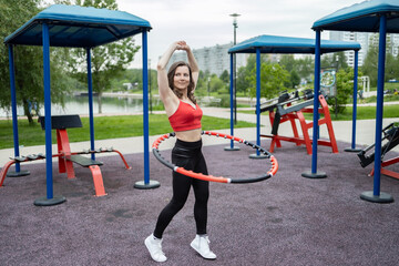 A girl in a red tank top and sweatpants does exercises and acrobatic tricks with a hula hoop on a...