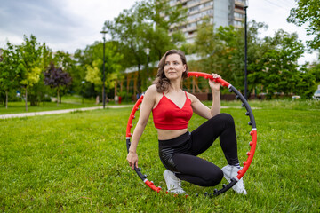A girl in a red tank top and sweatpants does exercises and acrobatic tricks with a hula hoop on a...
