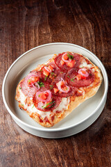 A close-up of a rectangular pizza slice with melted cheese, pepperoni, diced tomatoes, pickled onions, and fresh herbs, served on a white plate on a wooden table