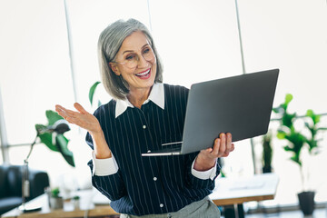 Smiling mature businesswoman using a laptop in a modern office setting, looking professional and...
