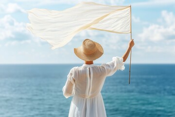 Woman in white dress and straw hat waves white flag by sea, standing with back to viewer in wind