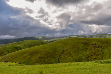 Rolling green hills and dense forests stretch towards a cloudy sky, showing rural landscape of...