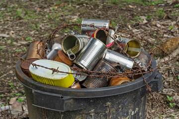 Rusty tin cans overflowing in a dirty trash bin wrapped with barbed wire, symbolizing pollution, danger, and environmental neglect in a rural area. Concept of waste and environmental hazard.