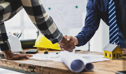Architects and engineers collaborate on construction projects. Two man are shaking hands over a construction plan on a wooden desk with a model house, a hardhat, a tape measure, and other tools.