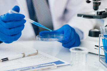Scientist analyzing samples with pipette in laboratory. Scientist wearing blue gloves is holding a pipette with blue liquid over a petri dish, conducting research in a laboratory setting.