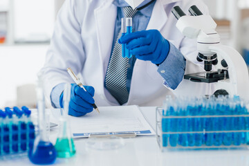 Scientist taking notes during experiment in laboratory. The lab table is filled with test tubes, beakers, and a microscope.
