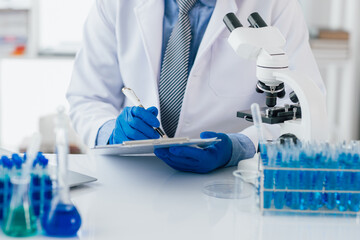 Scientist taking notes during experiment in laboratory. The lab table is filled with test tubes, beakers, and a microscope.