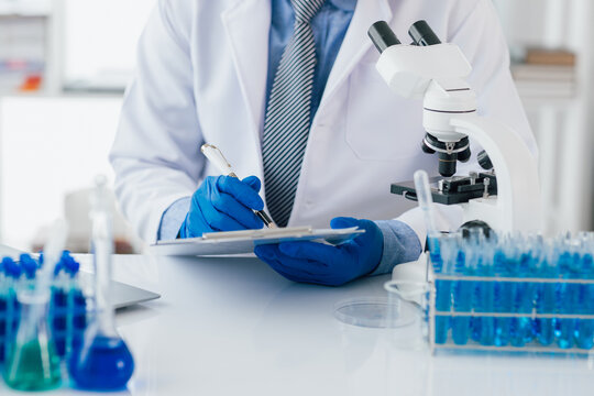 Scientist taking notes during experiment in laboratory. The lab table is filled with test tubes, beakers, and a microscope.