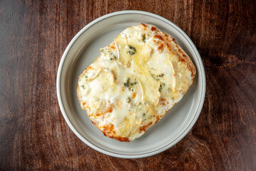 Golden, baked bread topped with melted cheese and aromatic herbs, served on a white plate over a rustic wooden table. The warm, crispy texture and gooey cheese