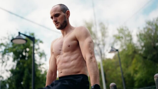 A muscular man performs dips on parallel bars in an urban park setting. He is shirtless and focused on his workout. This video represents fitness, strength training, and healthy lifestyle.