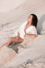 brunette girl in white boho style dress and silver jewelries is lying casual and looking apart on the sand dunes career background in desert at sunny day. rustic style. lifestyle concept, free space