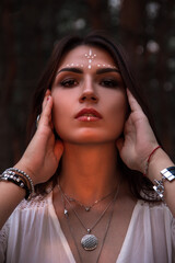 young brunette girl with face painting in white boho dress and jewelry is standing like magic shaman in fairytale with hands near head and looking straight on forest background in sun lights at sunset