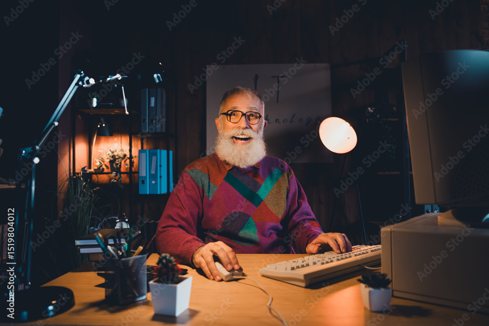 Poster smiling elderly man in a colorful sweater working on a desktop computer in a cozy home office with w
