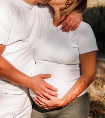 close up male hand on the pregnant belly. pregnant couple in white t-shirts hugs outdoors at sunny day. male and female hands with mehendi on pregnant girl belly. pregnant concept, free space