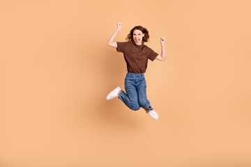 Excited young woman in casual jeans and brown t-shirt jumping joyfully against a beige background