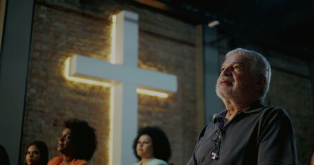 Elderly man in church looking at illuminated cross, expression of faith and devotion during prayer,...