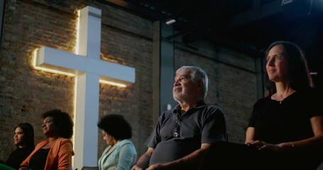 Diverse group of people seated in church with illuminated cross in the background, expressing spirituality and devotion during prayer, unity and connection in religious gathering