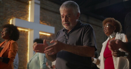 Elderly man clapping during church worship, illuminated cross in background, diverse group expressing faith, unity, and devotion in religious indoor gathering