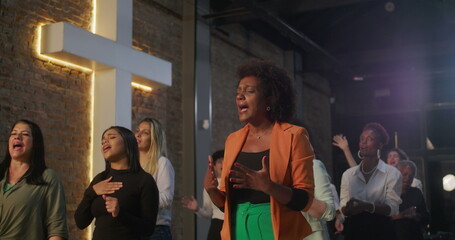 Women singing and worshiping together in diverse church community, standing near illuminated cross,...