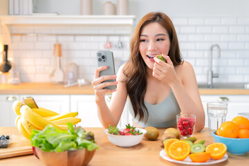 cheerful Asian woman in sporty casual wear enjoys a healthy fruit smoothie in her modern kitchen after a home workout, smiling while video calling a friend, embracing an easy, balanced lifestyle focus