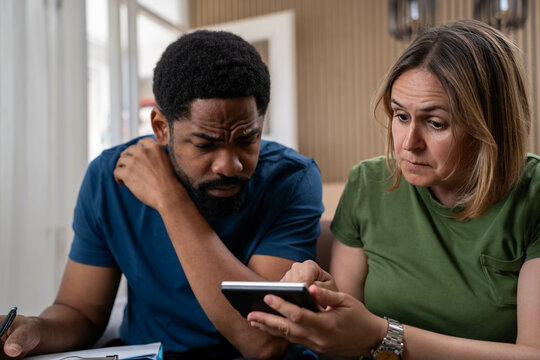 Worried Black man and White woman sitting in living room, stressed about rising living costs, bills, rent, mortgages, and personal finance challenges during inflation crisis.