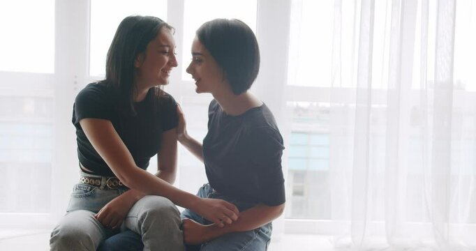 Low angle handheld shot of young lesbian couple smiling and touching hands gently while sitting on sofa against window in light living room at home. Two women sharing love and support holding hands.