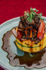 Causa rellena de lomo saltado, en plato claro, sobre mesa de mantel color rojo.