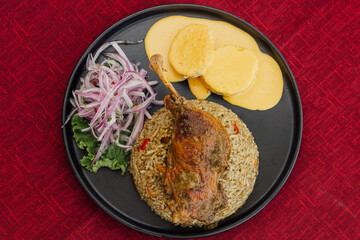 Plano cenital del arroz con pato, acompañado de papa a la huancaína, más ensalada de cebolla, en plato oscuro, sobre una mesa de mantel rojo.