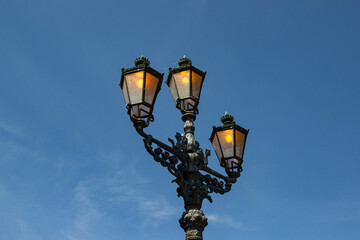 Vintage three-lantern streetlight shaped like a trident, lit during daytime against a clear blue...