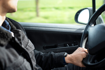 Close-up of a man's hands gripping the steering wheel while driving a car. Green foliage is visible through the window, suggesting movement and a natural outdoor environment. 