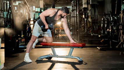 Strong man performing one-arm dumbbell row on gym bench with focus and determination under dramatic...