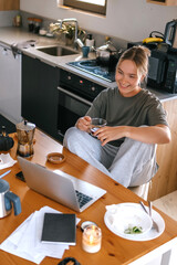  Gen Z woman working from home at cozy kitchen table with laptop and phone. Digital nomad...