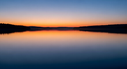 Fototapeta premium Serene sunset reflections on a calm lake with silhouette of forested shoreline and horizon