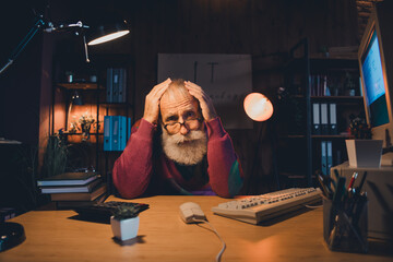 Elderly man working late on computer in cozy home office setting with focused expression,...