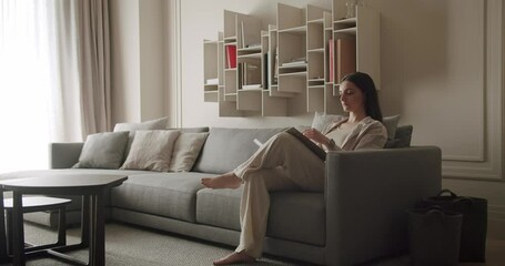 Woman sitting on a comfortable sofa reading a book in a modern, cozy living room with stylish shelving and minimalist design. Woman Reading Book On Sofa In Modern Living Room With Stylish Shelving - Powered by Adobe
