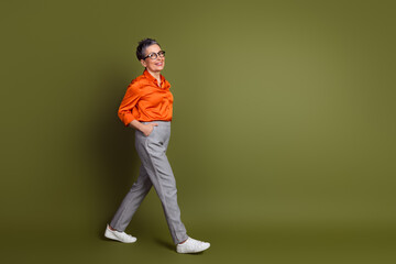 Confident mature woman wearing stylish orange blouse and gray trousers walking against khaki...