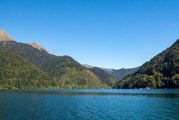 Lake Ritsa surrounded by mountain peaks. Boats sail on smooth turquoise water. Mountain slopes are covered with dense forest. Sunny autumn day