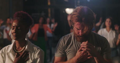 Congregation in prayer during spiritual gathering, hands on hearts, diverse group expressing faith and devotion, solemn worship moment with illuminated cross in background