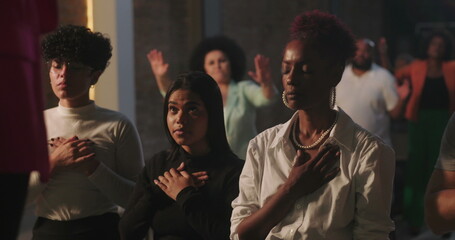 Congregation in prayer during spiritual gathering, hands on hearts, diverse group expressing faith and devotion, solemn worship moment with illuminated cross in background